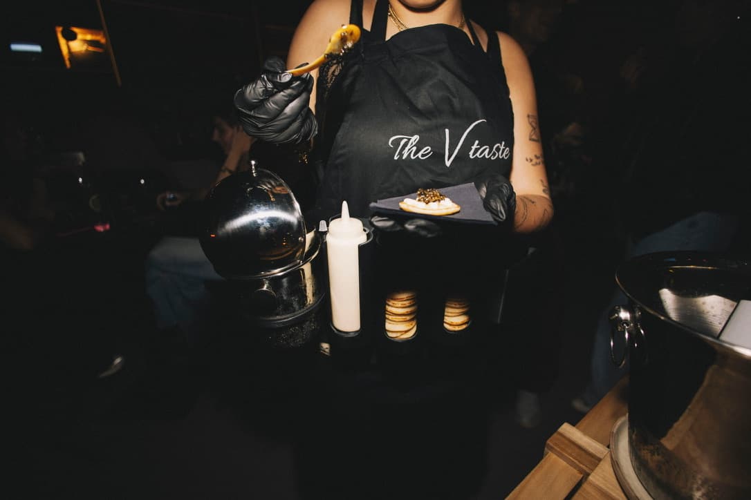 A TheVtaste chef in a branded apron plating a caviar blini tableside