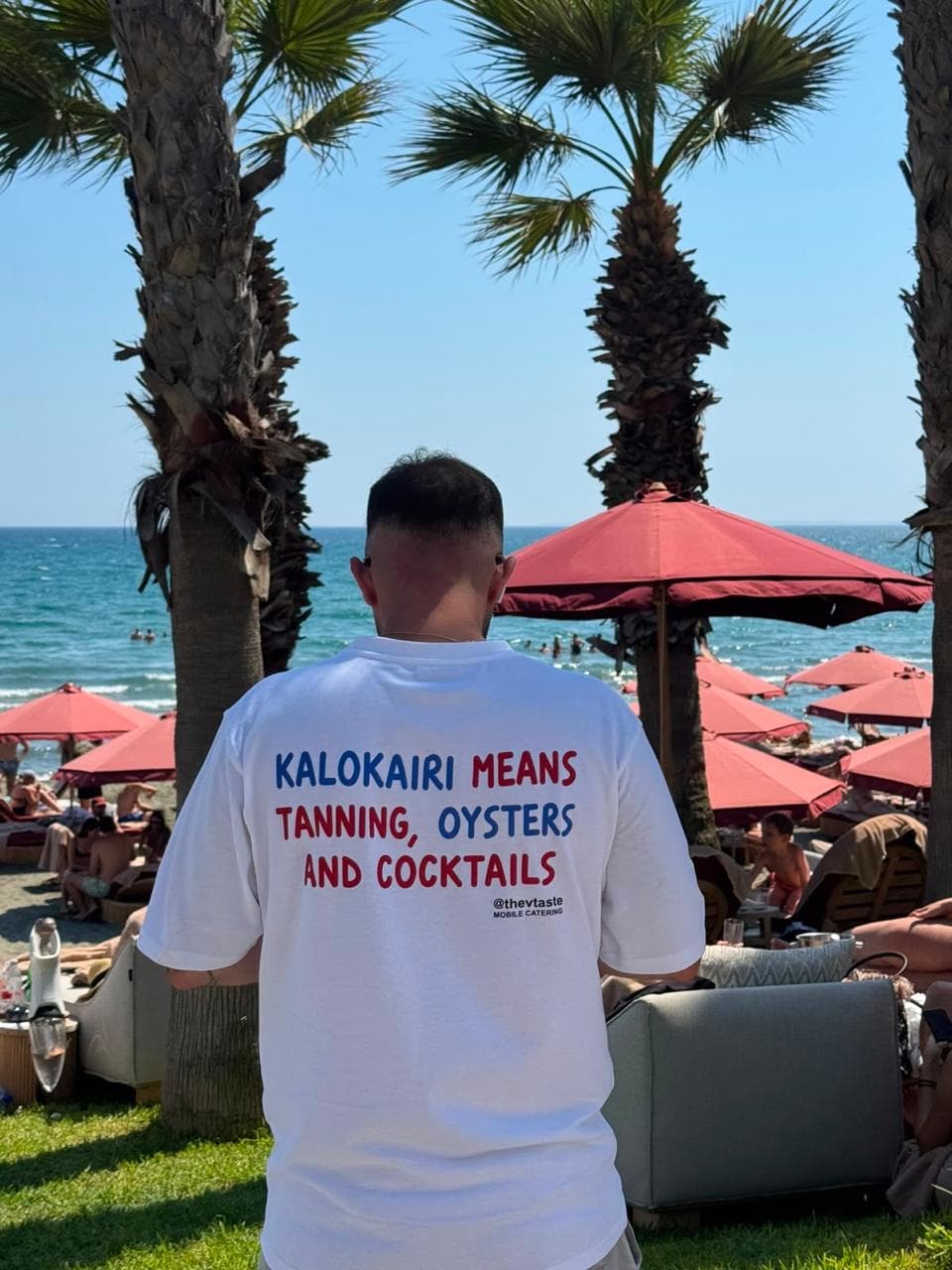 A TheVtaste crew member on a beach in Cyprus, wearing a branded t-shirt reading 'Kalokairi means tanning, oysters and cocktails'